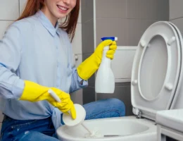 Une femme qui nettoie les toilettes avec des gants jaunes et un pchitt et une brosse à toilettes