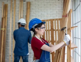 une femme en casque bleue et en salopette qui tape contre un mur en bois avec un marteau