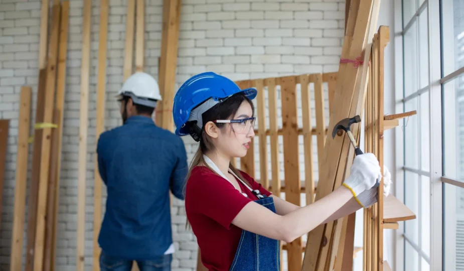 une femme en casque bleue et en salopette qui tape contre un mur en bois avec un marteau