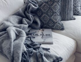 A stylish living room featuring a white sofa with patterned pillows, a blanket, and a fashion book.