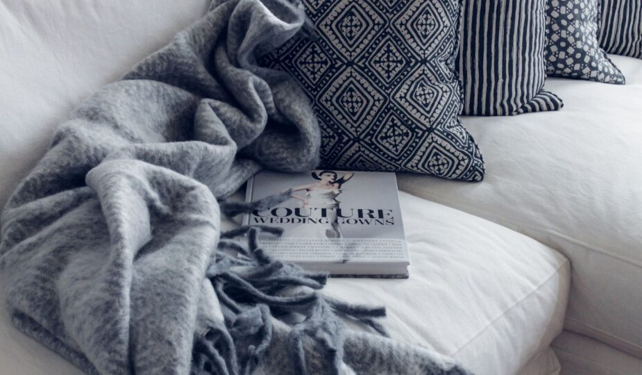 A stylish living room featuring a white sofa with patterned pillows, a blanket, and a fashion book.