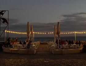 Cozy outdoor dining area by the water at twilight, adorned with glowing string lights.