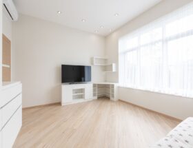 Modern TV set on cupboard placed near empty table in spacious room with big window and white cabinets in apartment