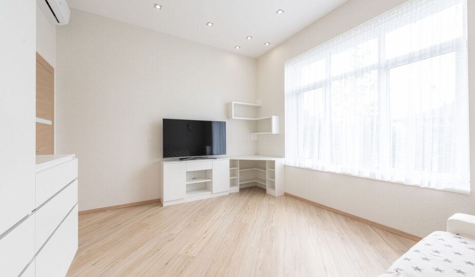 Modern TV set on cupboard placed near empty table in spacious room with big window and white cabinets in apartment