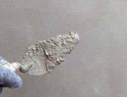 Hand wearing glove holds plaster trowel against smooth wall, depicting construction work.