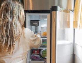 Back view of a blonde woman retrieving food from a refrigerator in a bright kitchen.