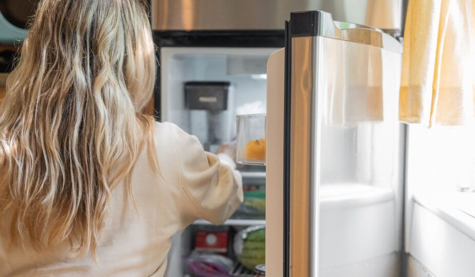 Back view of a blonde woman retrieving food from a refrigerator in a bright kitchen.