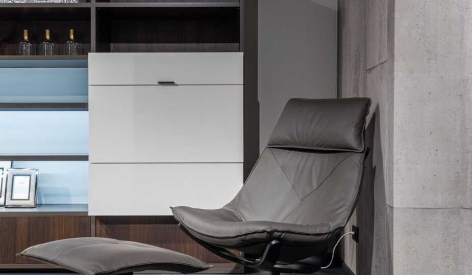 Comfortable leather chair with footrest placed on carpet near cabinet with shelves in modern flat