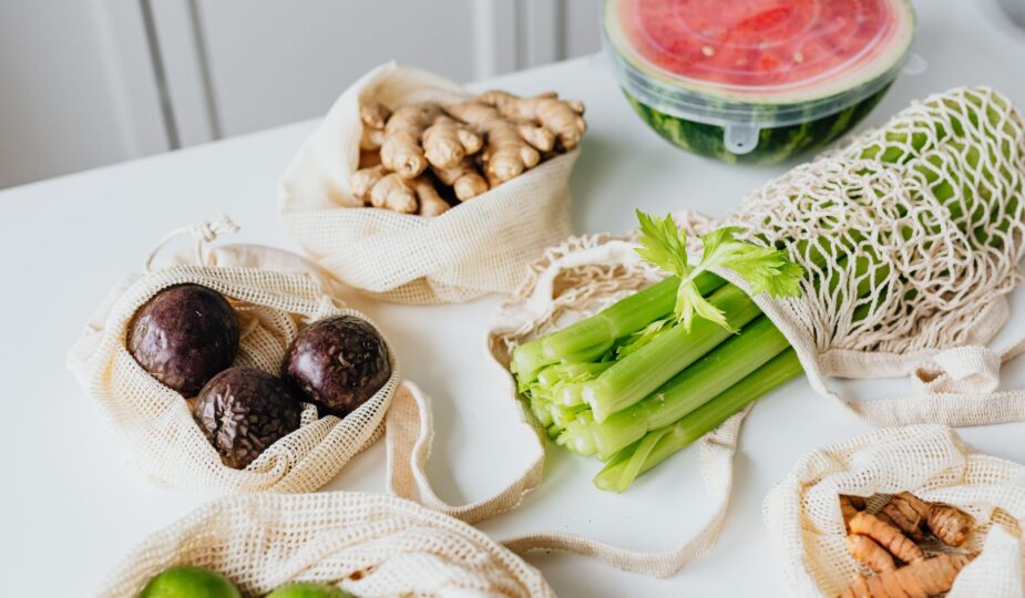 Assortment of fresh vegetables and fruits in eco-friendly net bags on a white table.